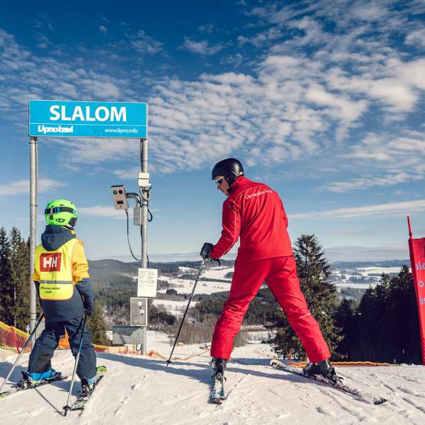 Malý lyžař s instruktorem se chystá sjet sjezdovku s měřeným slalomem